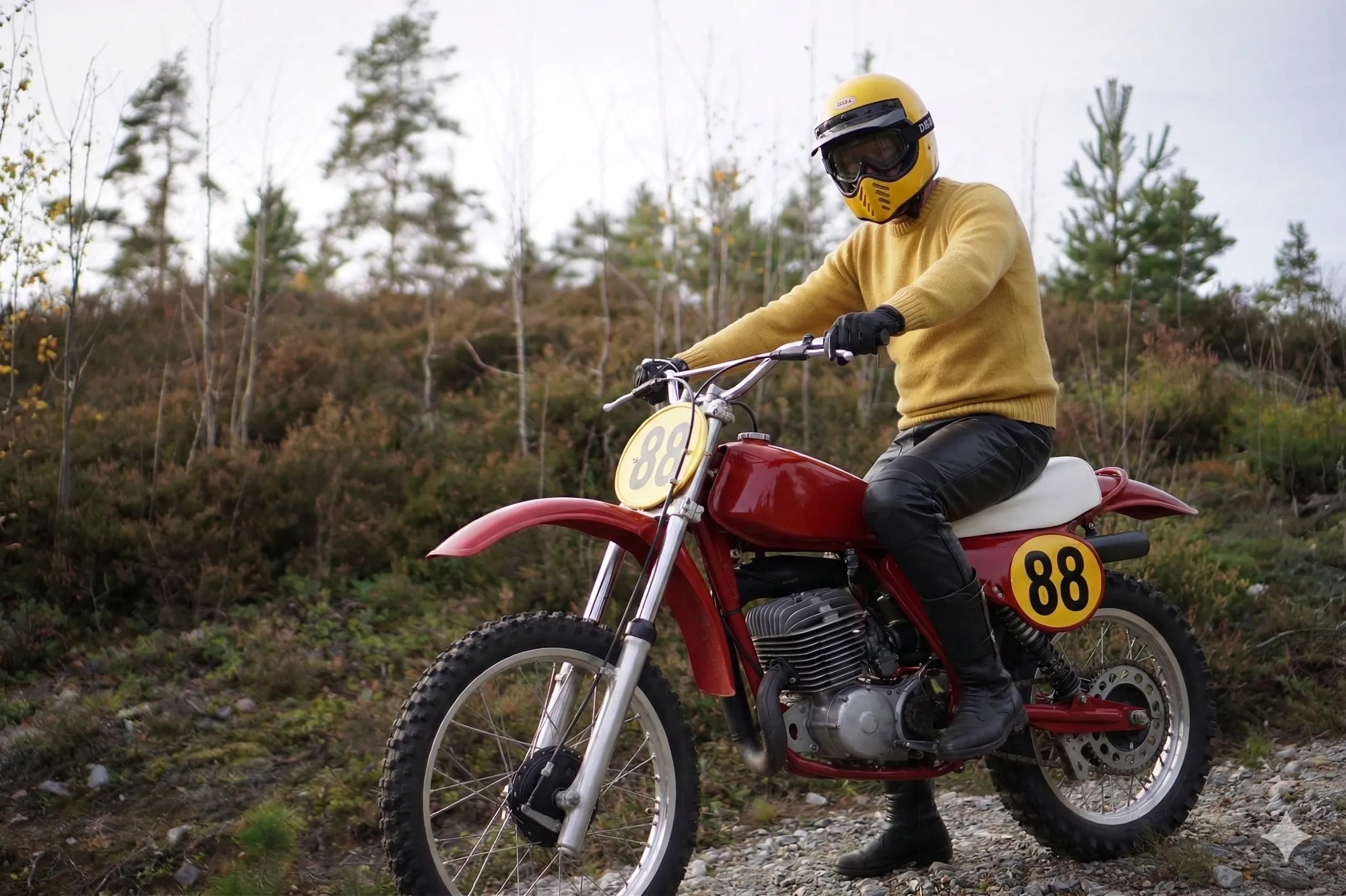 A person wearing a yellow sweater and yellow helmet sits on a classic red motocross bike in a wooded area.
